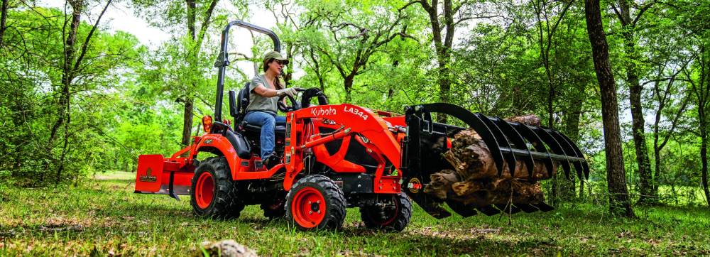 Kubota BX Series tractor with a grapple attachment loading logs on a wooded property